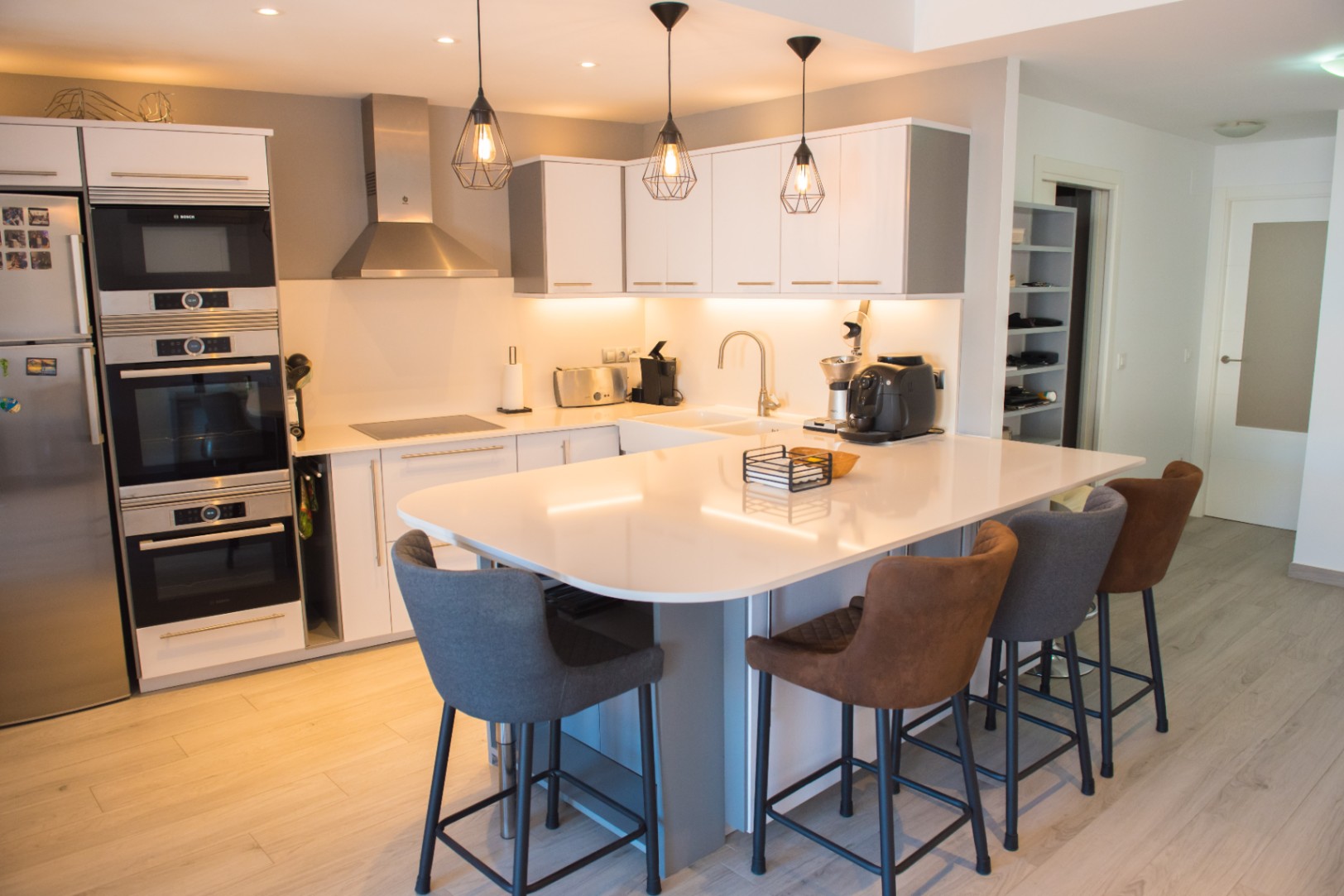 Cuisine moderne avec îlot central blanc, tabourets élégants, électroménagers Bosch encastrés et luminaires suspendus de style industriel.