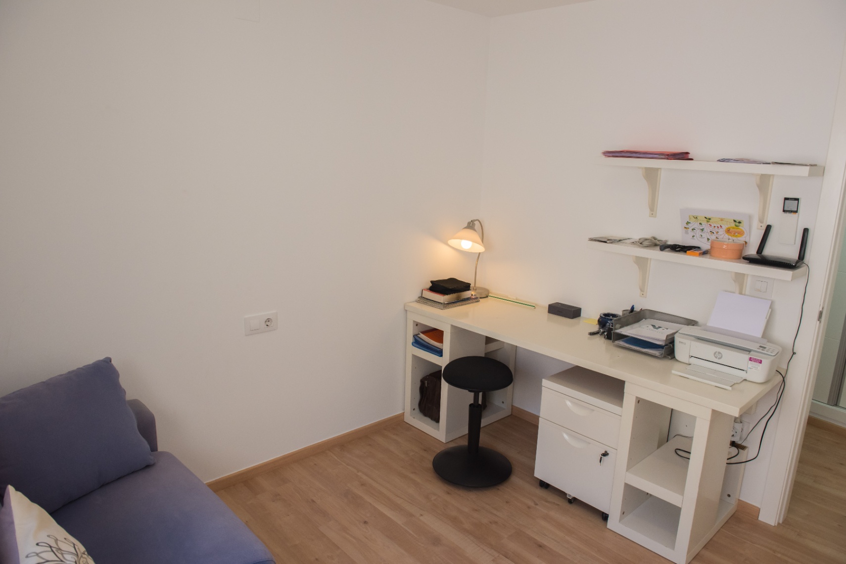 Espace bureau fonctionnel comprenant un bureau blanc avec rangements intégrés, un tabouret noir réglable et des étagères murales sur parquet clair.