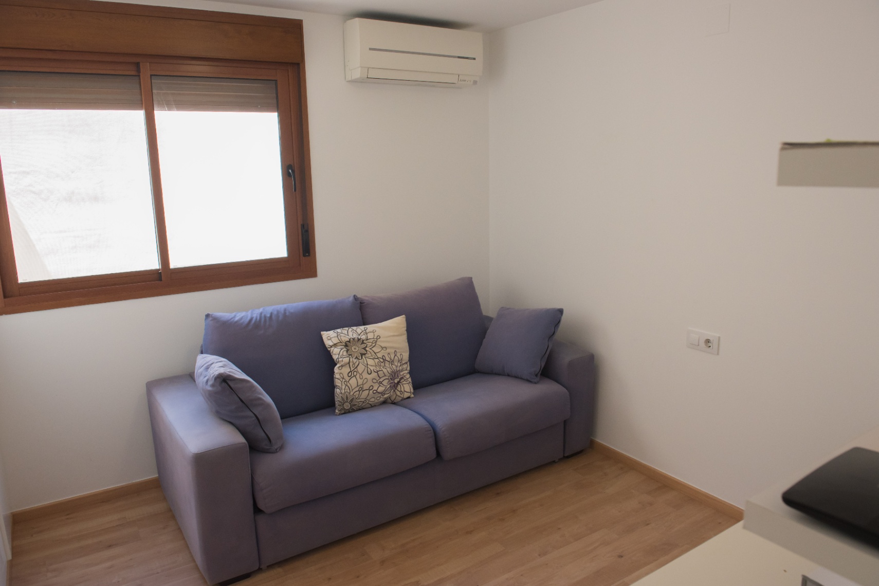 Espace de vie comprenant un canapé bleu, un parquet en bois clair et une unité de climatisation murale près d'une fenêtre en bois.