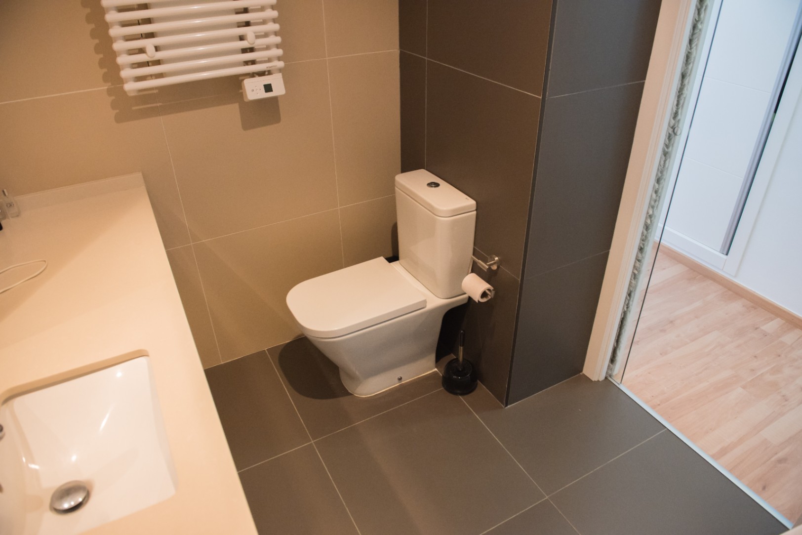 Salle de bain moderne avec toilettes en céramique blanche, carrelage gris foncé et sèche-serviettes mural. Plan de toilette crème avec vasque intégrée.