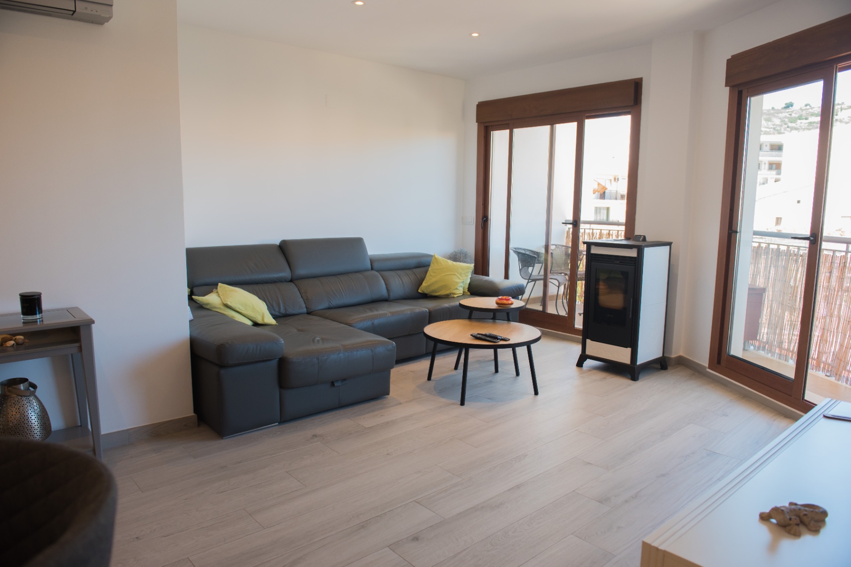 Salon contemporain avec canapé d'angle en cuir foncé, tables basses gigognes en bois et poêle à granulés. Les baies vitrées en bois apportent une luminosité naturelle abondante.