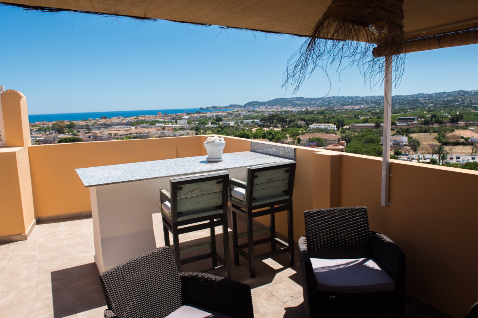 Terrasse comprenant une table de bar en granit avec tabourets hauts et fauteuils en osier, offrant une vue panoramique sur la ville côtière et la mer.