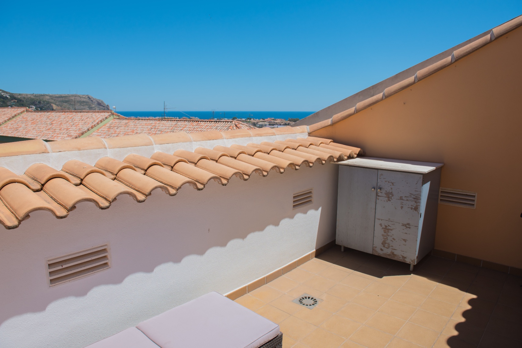 Terrasse de toit avec carrelage au sol, muret en tuiles romanes et vue lointaine sur la mer. Comprend un meuble de rangement extérieur fonctionnel.