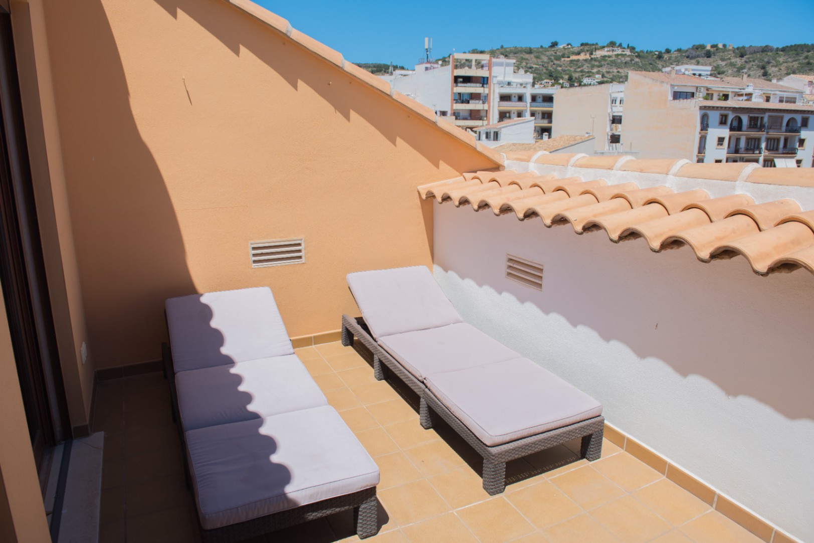 Terrasse de toit privée avec deux chaises longues en rotin gris, sol en terre cuite et vue sur la ville et les collines environnantes.