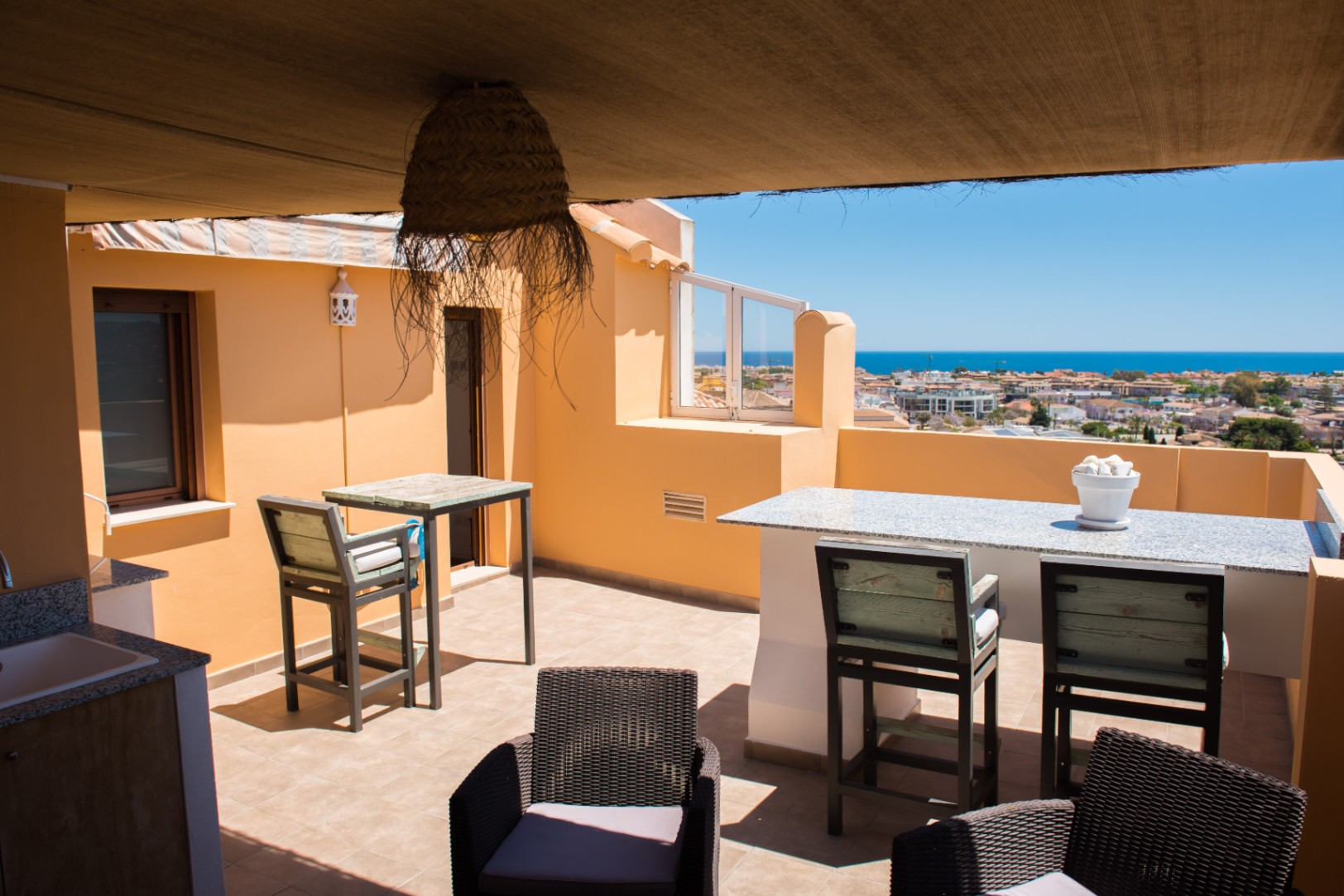 Terrasse sur le toit avec bar en granit, mobilier de jardin et vue panoramique sur la mer. Comprend un espace ombragé avec luminaire en osier et sol carrelé.