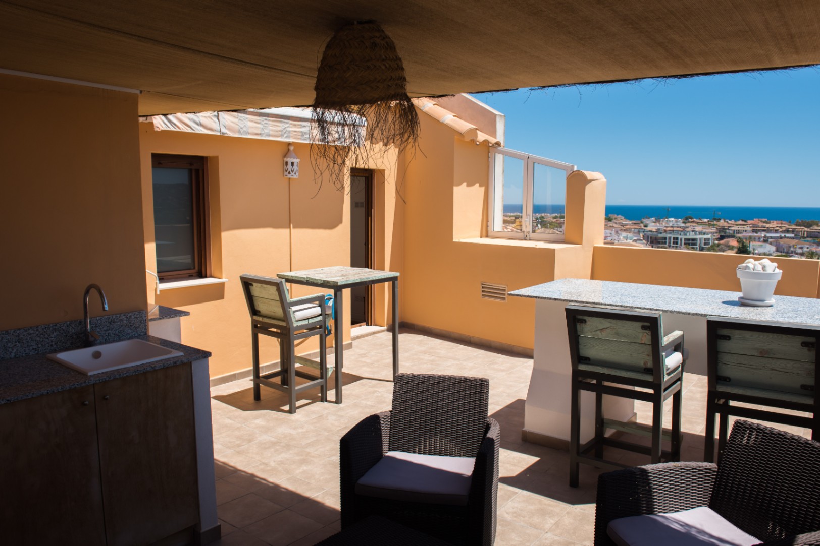 Terrasse sur le toit avec cuisine d'été en granit, coin bar et fauteuils en rotin, offrant une vue dégagée sur la mer Méditerranée et le paysage urbain.
