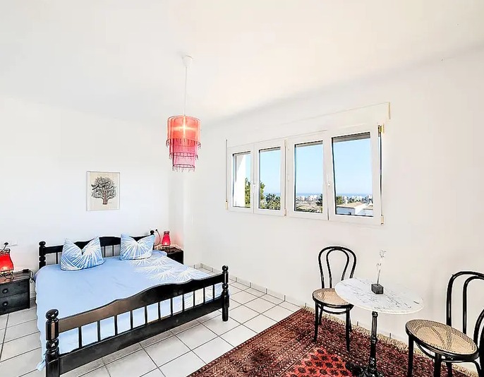 Chambre lumineuse avec carrelage blanc, lit en bois sombre et coin salon avec table en marbre. Les fenêtres offrent une vue dégagée sur le paysage et la mer.