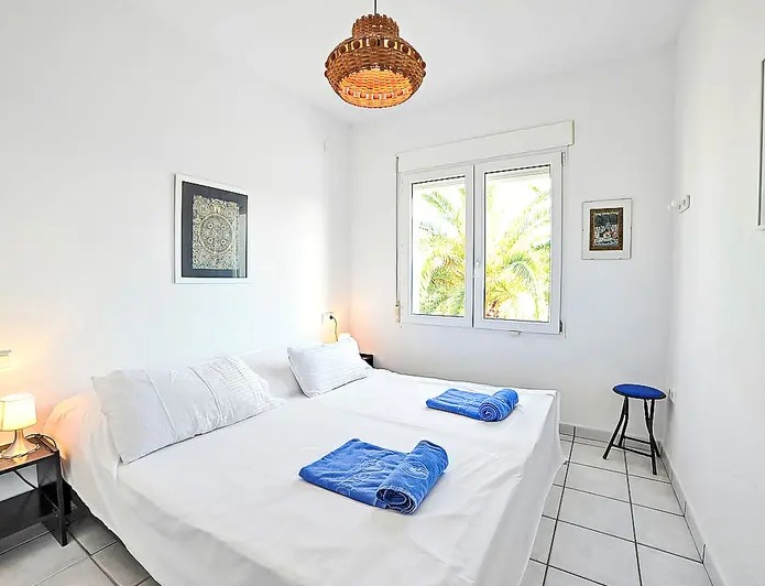 Chambre lumineuse avec murs blancs, sol en carrelage et fenêtre offrant une vue sur les palmiers. Elle dispose d'une suspension en osier et de lits jumeaux.