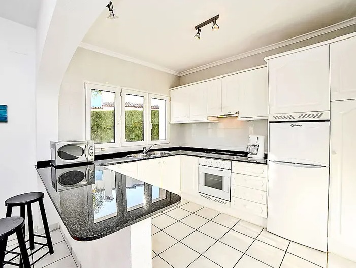 Cuisine équipée avec meubles blancs, plan de travail en granit noir et bar. Comprend des appareils intégrés, un sol en carrelage et une fenêtre offrant une luminosité naturelle.