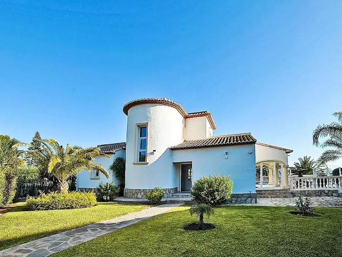 Façade extérieure de la villa avec une tour circulaire, des murs blancs et un toit en terre cuite. Le jardin soigné comprend une pelouse et des palmiers.