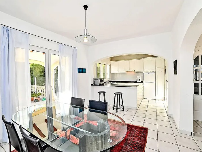 Salle à manger lumineuse avec table en verre, s'ouvrant sur une cuisine blanche équipée avec bar via une arche méditerranéenne élégante.