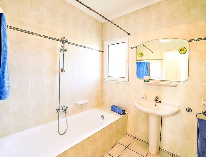 Salle de bain fonctionnelle avec baignoire, lavabo sur colonne et murs carrelés dans les tons crème avec une frise décorative.