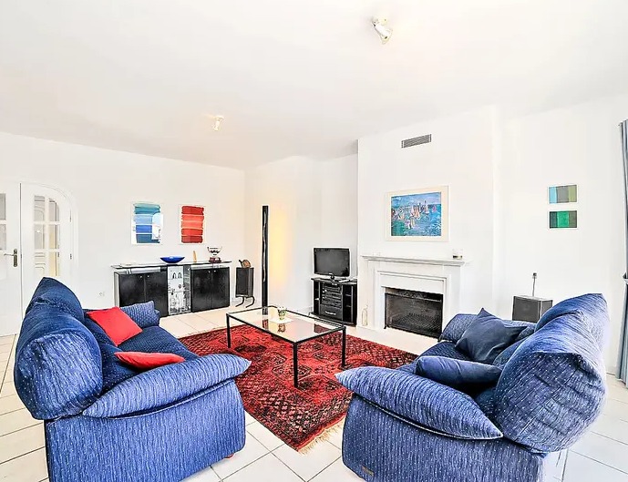 Salon lumineux avec cheminée blanche, canapés bleus, tapis oriental rouge et carrelage clair offrant une sensation d'espace.