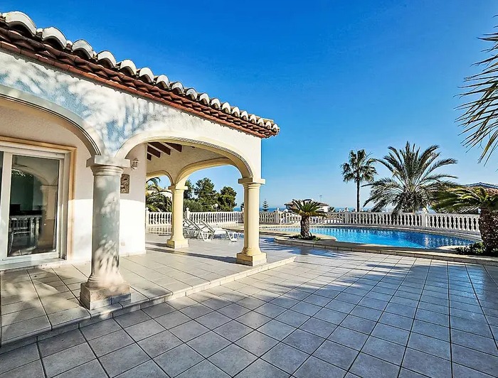 Terrasse extérieure avec naya voûtée, colonnes en pierre, carrelage clair et piscine privée entourée de palmiers avec vue dégagée.