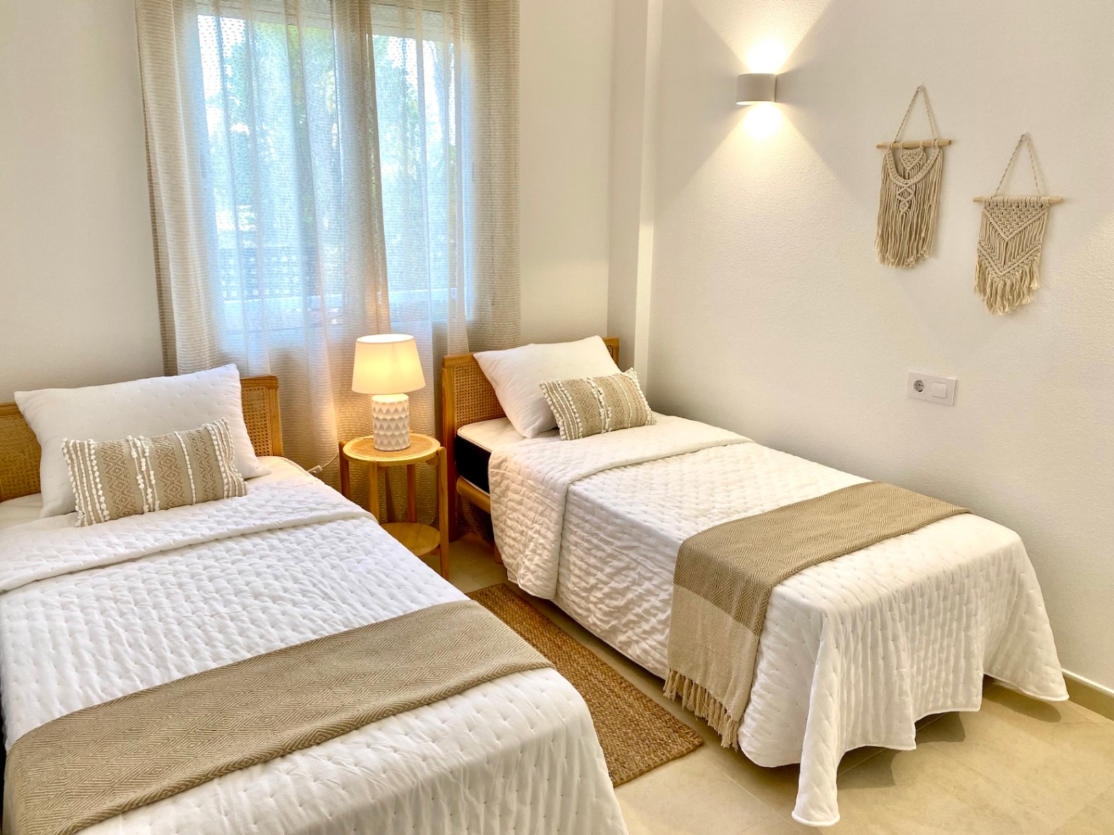 Chambre d'amis lumineuse avec deux lits simples, têtes de lit en osier, textiles naturels, décorations en macramé et éclairage tamisé par des appliques et lampes.