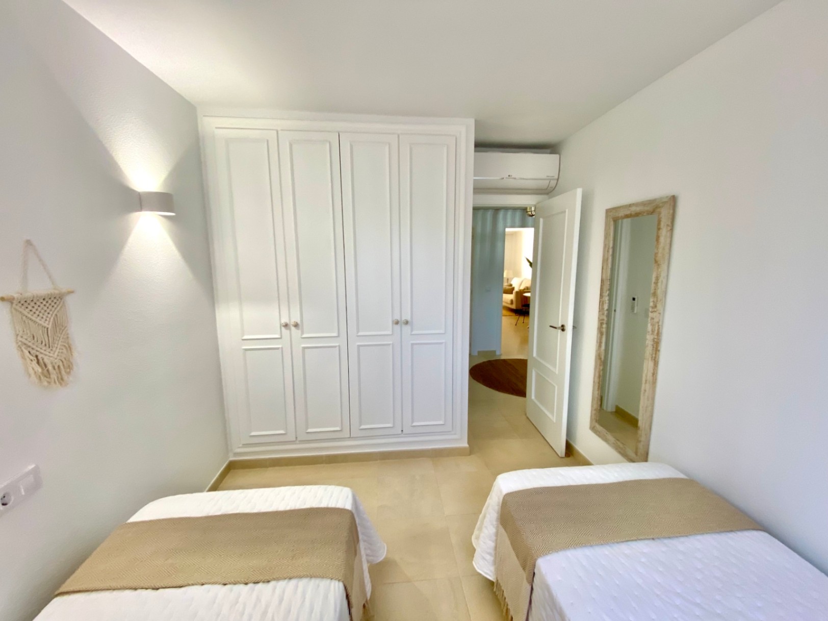 Chambre lumineuse avec lits jumeaux, une grande armoire encastrée blanche à panneaux et un miroir au cadre en bois rustique sur murs blancs.
