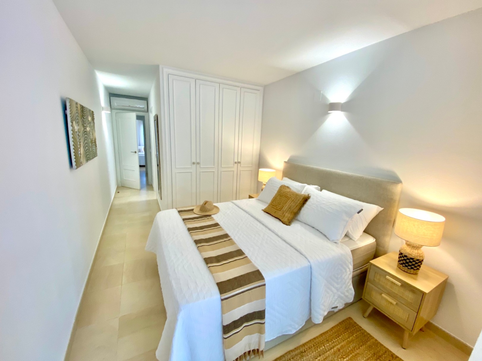 Chambre moderne avec placards intégrés blancs, tête de lit rembourrée beige et tables de chevet en bois avec éclairage tamisé sur sol carrelé.