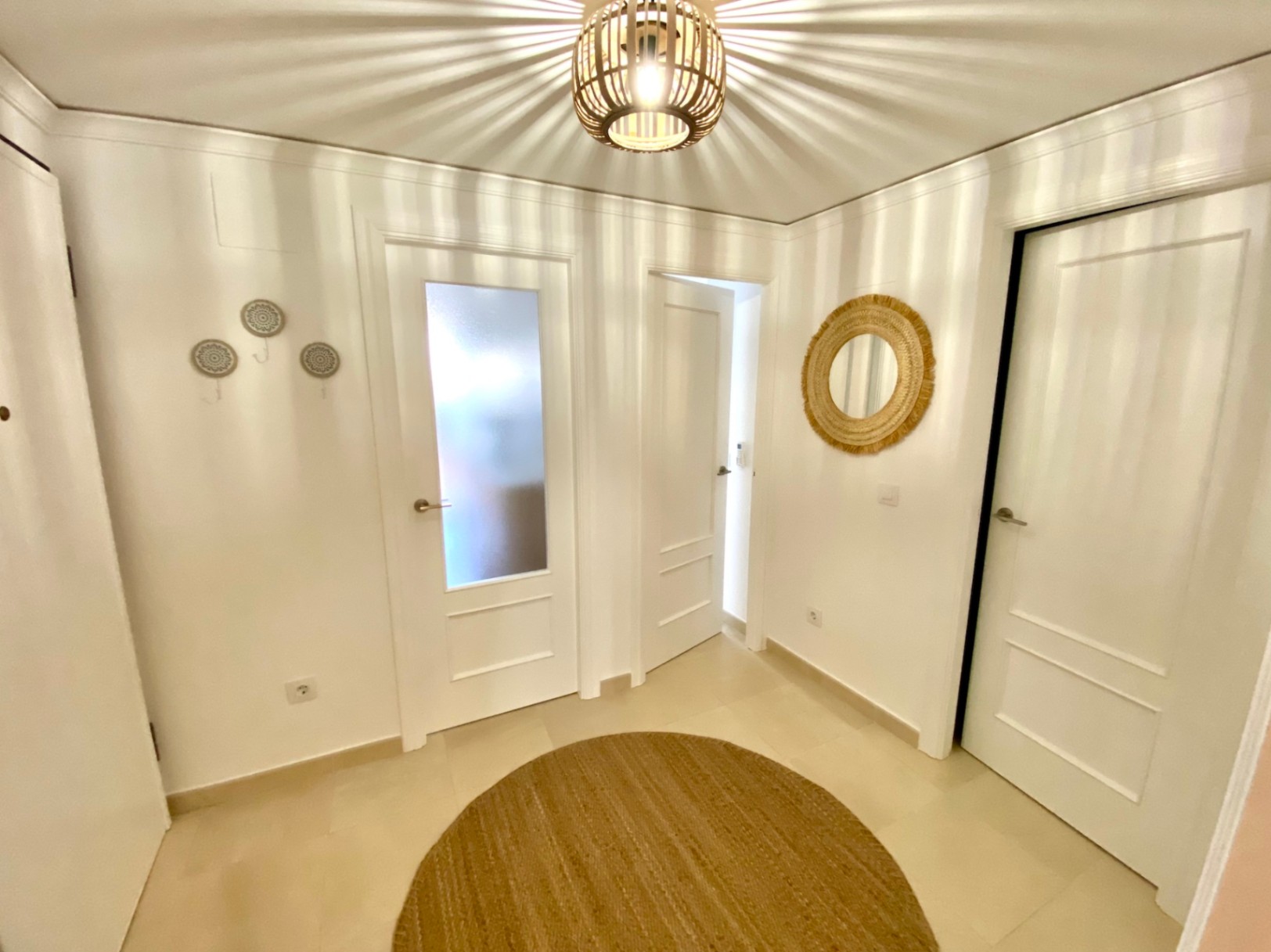 Couloir avec portes blanches, tapis rond en jute et suspension en bois projetant des ombres radiales décoratives sur les murs et le plafond.