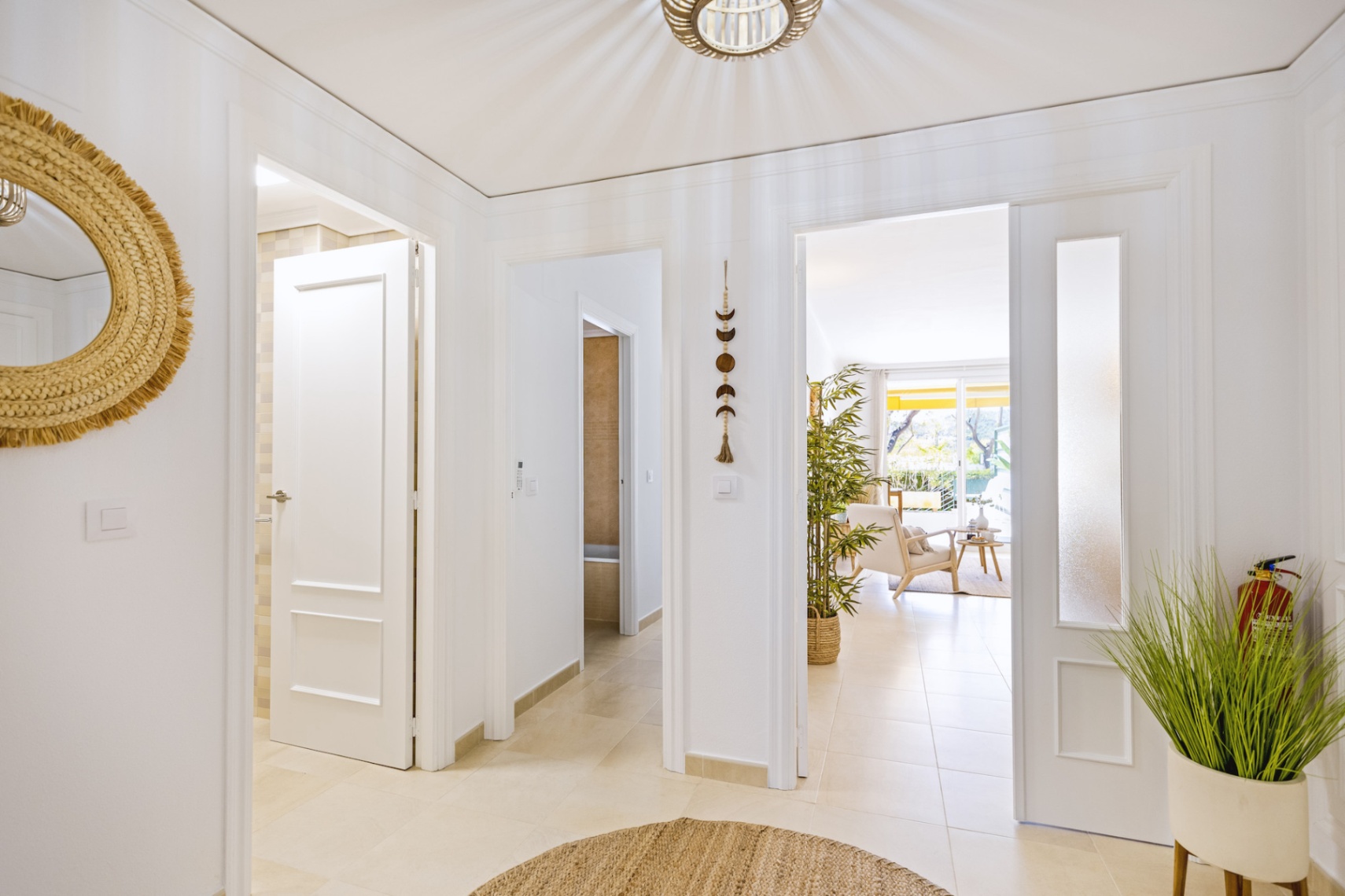 Couloir de distribution lumineux avec carrelage clair, portes blanches moulurées et miroir en osier, menant vers le salon ensoleillé.