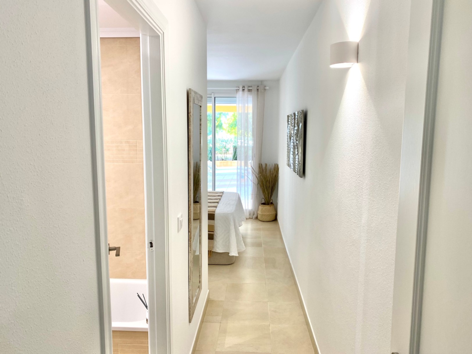 Couloir lumineux avec carrelage beige menant à une chambre avec accès terrasse. Salle de bain attenante avec faïence murale de ton neutre et baignoire.