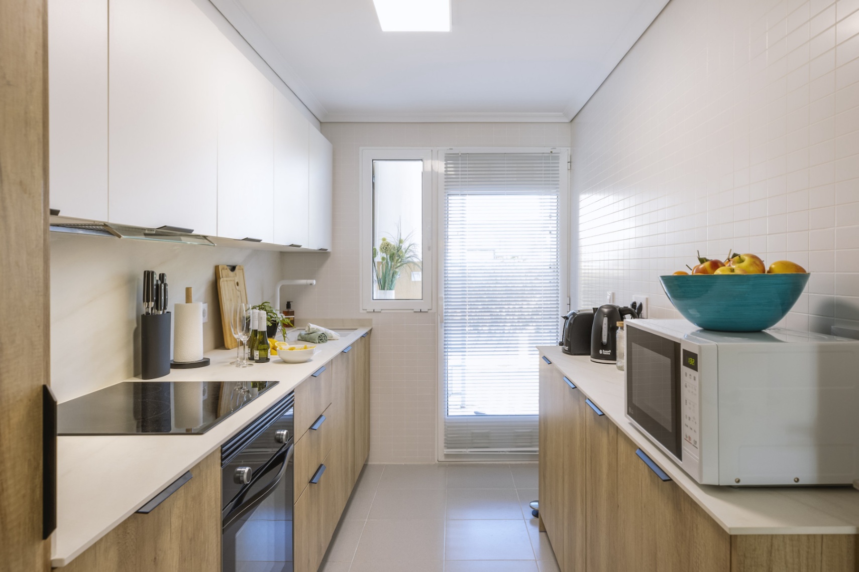 Cuisine en couloir moderne avec meubles bas en bois, placards hauts blancs, électroménager intégré et fenêtre apportant une lumière naturelle.