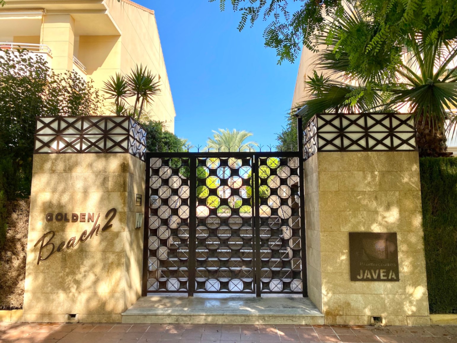 Entrée du Golden Beach 2 à Javea, avec un portail métallique aux motifs circulaires encadré par des piliers en pierre et des palmiers.