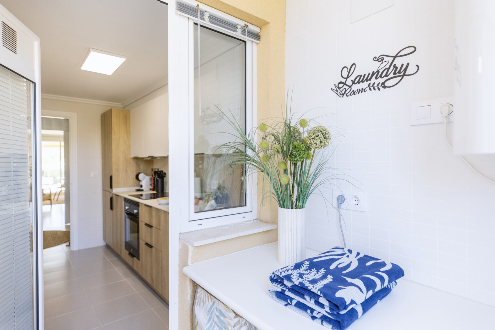 Espace buanderie fonctionnel avec plan de travail blanc, situé à côté d'une cuisine moderne avec des meubles en bois et de l'électroménager encastré.