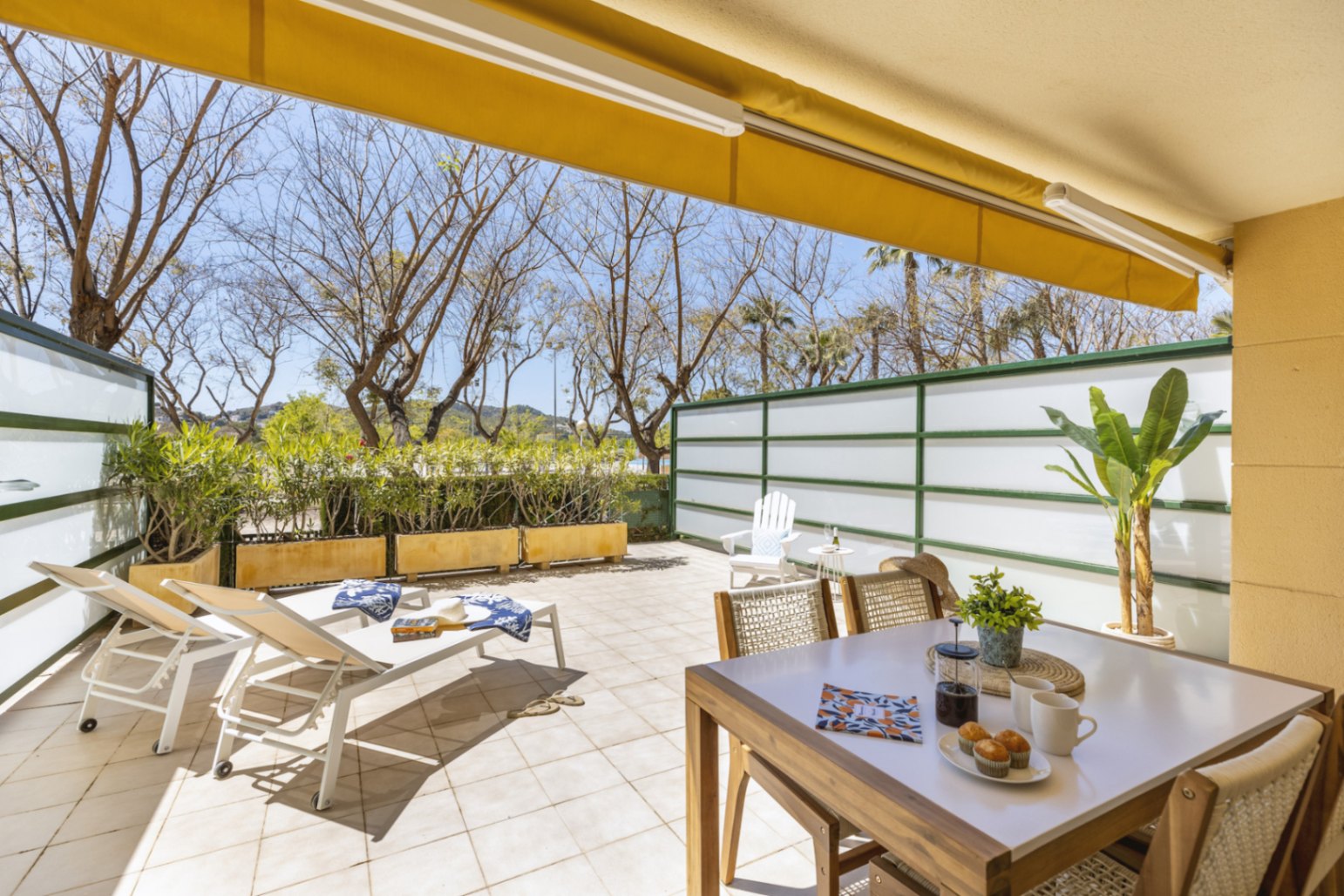 Grande terrasse carrelée avec store banne jaune, coin repas extérieur et chaises longues blanches, entourée de parois d'intimité et d'arbres matures.
