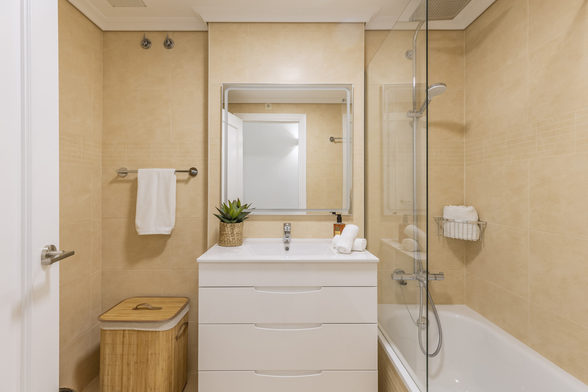 Salle de bain moderne avec carrelage beige, meuble vasque blanc à tiroirs, miroir rétroéclairé et baignoire avec paroi de douche en verre.