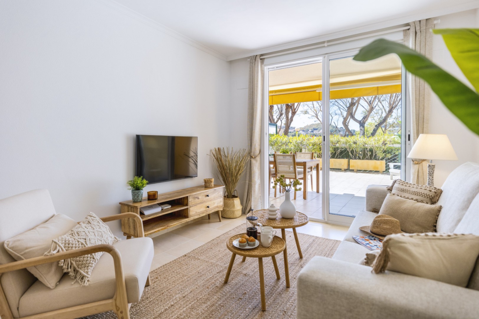 Salon lumineux avec mobilier en bois, tapis en jute et tables en rotin, s'ouvrant sur une terrasse ensoleillée par de larges baies vitrées.