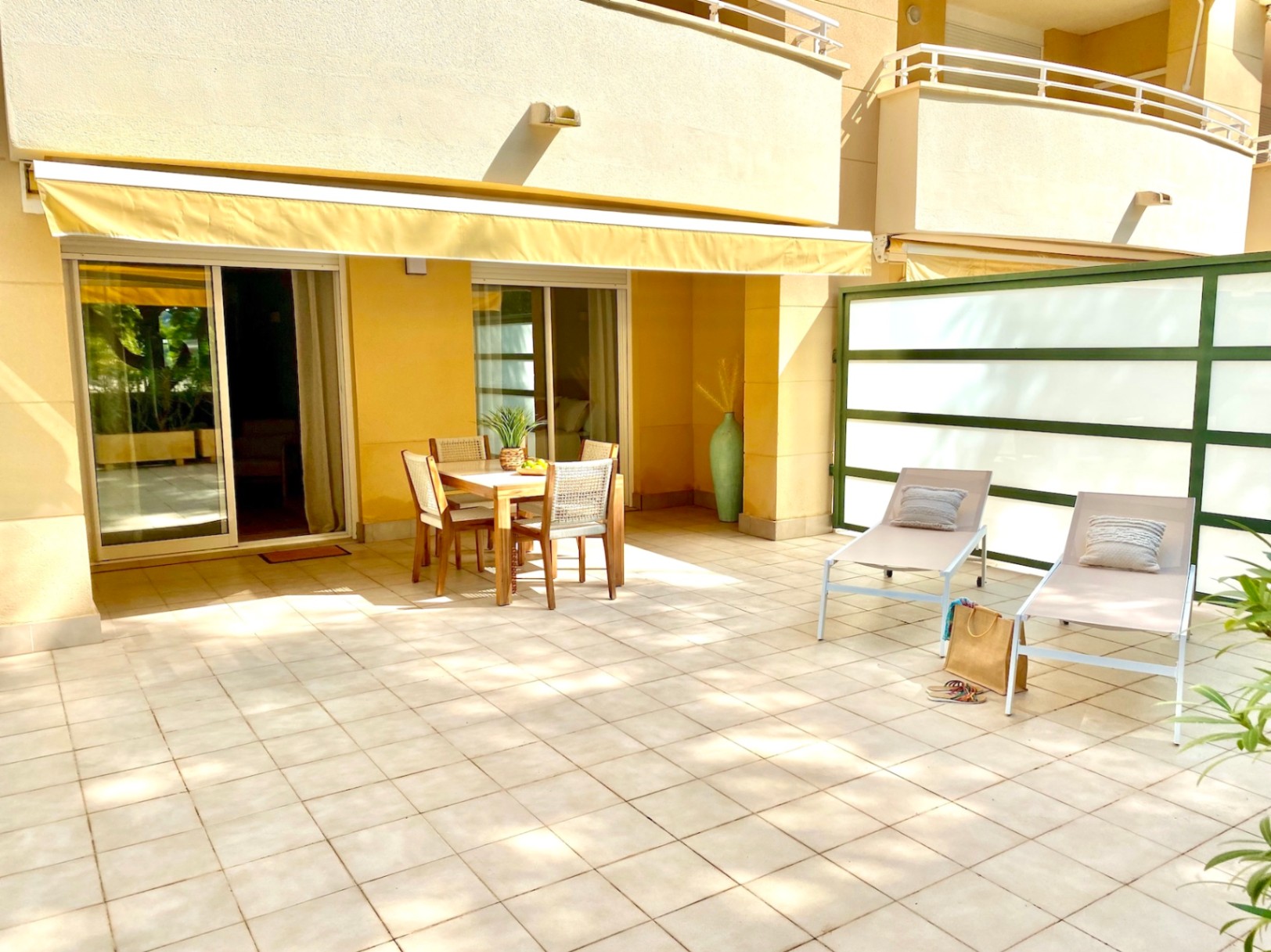 Vaste terrasse carrelée comprenant un ensemble repas en bois sous un store banne jaune et deux bains de soleil bordés d'un écran d'intimité.