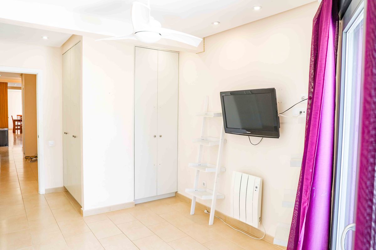 Chambre lumineuse avec placards intégrés blancs, télévision murale et étagère échelle. Équipée d'un ventilateur de plafond, de spots encastrés et d'un sol carrelé.