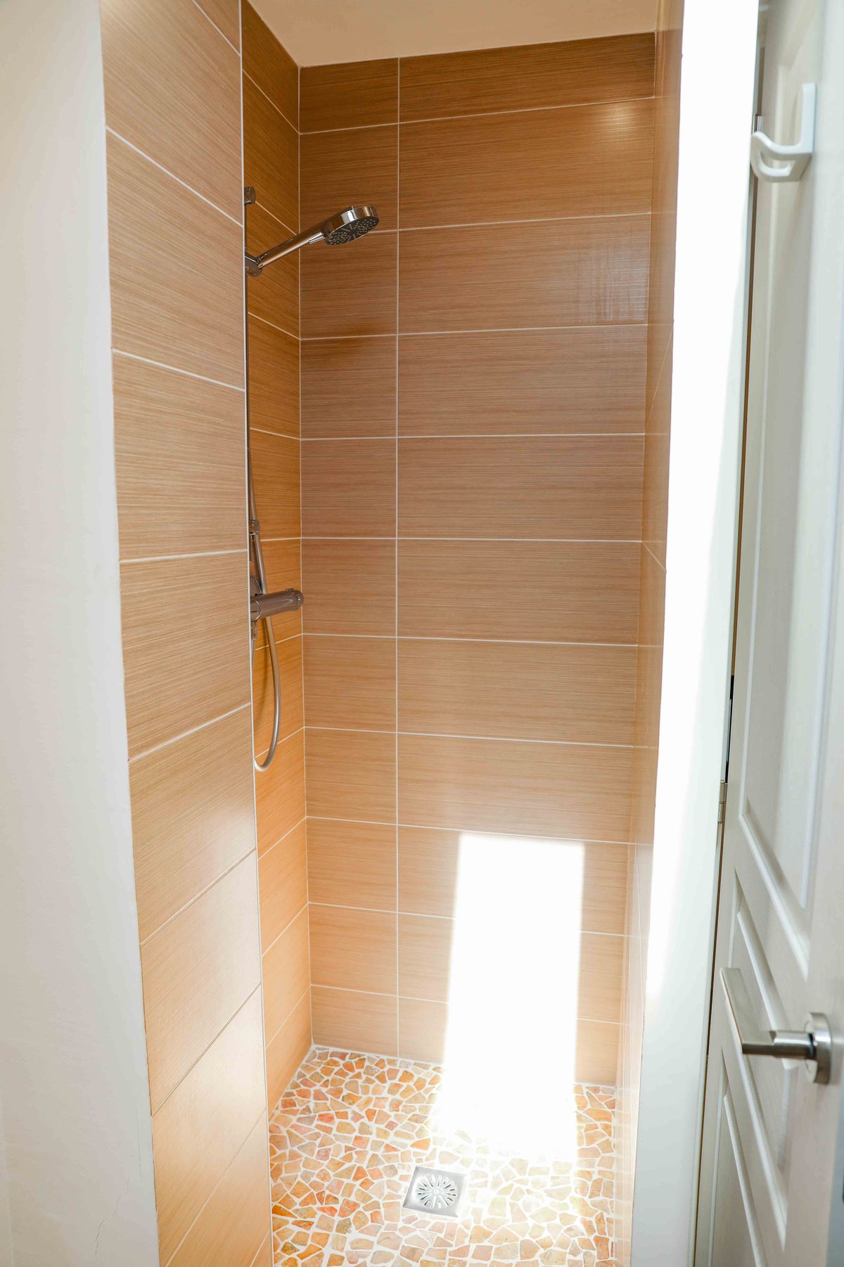 Douche à l'italienne avec carrelage mural horizontal effet bois et sol en mosaïque de galets. Équipée d'une pomme de douche chromée et de lumière naturelle.