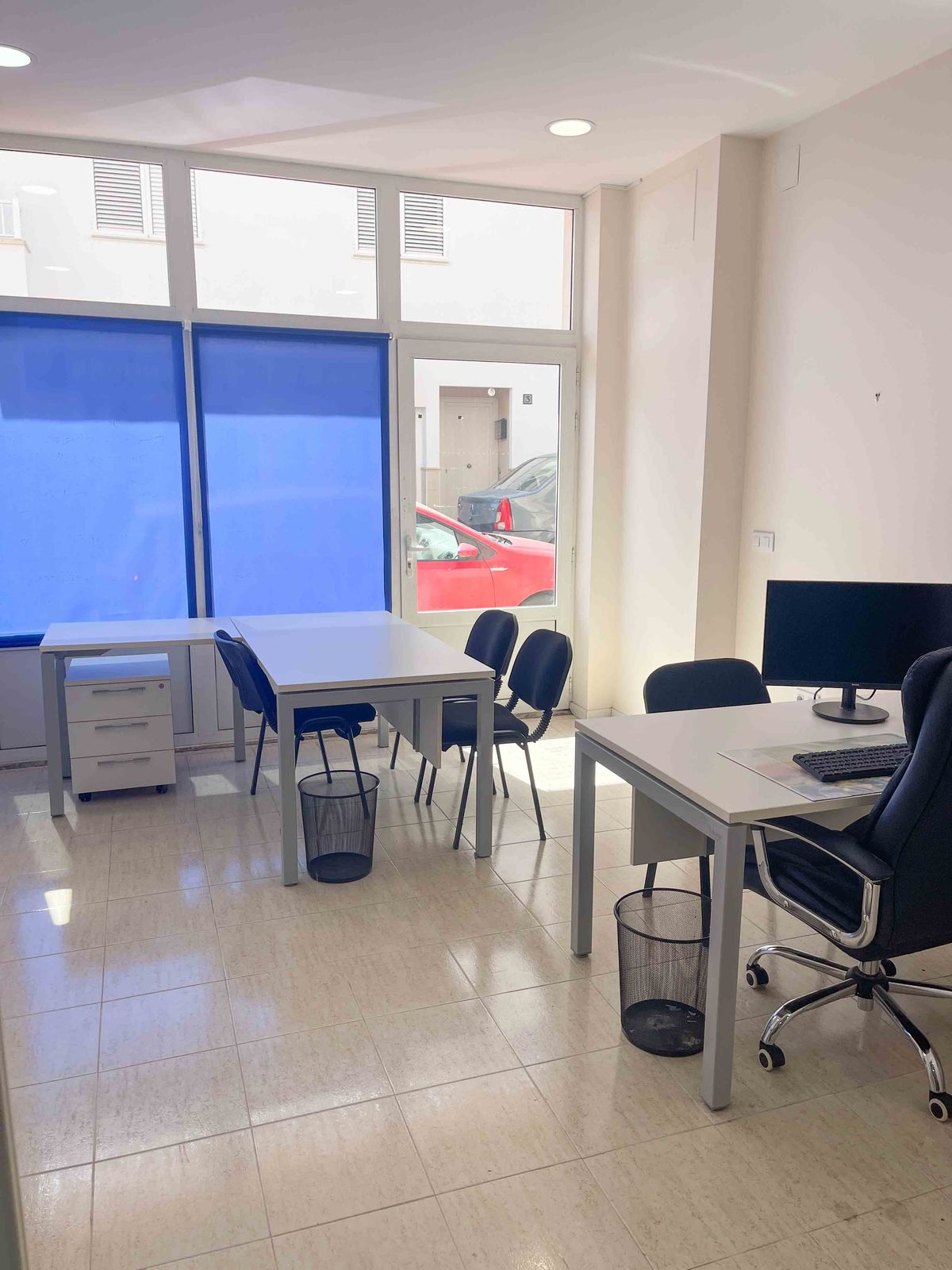 Espace de bureau lumineux avec postes de travail blancs, chaises ergonomiques noires et sol carrelé. Grandes fenêtres avec stores bleus et accès direct à la rue.