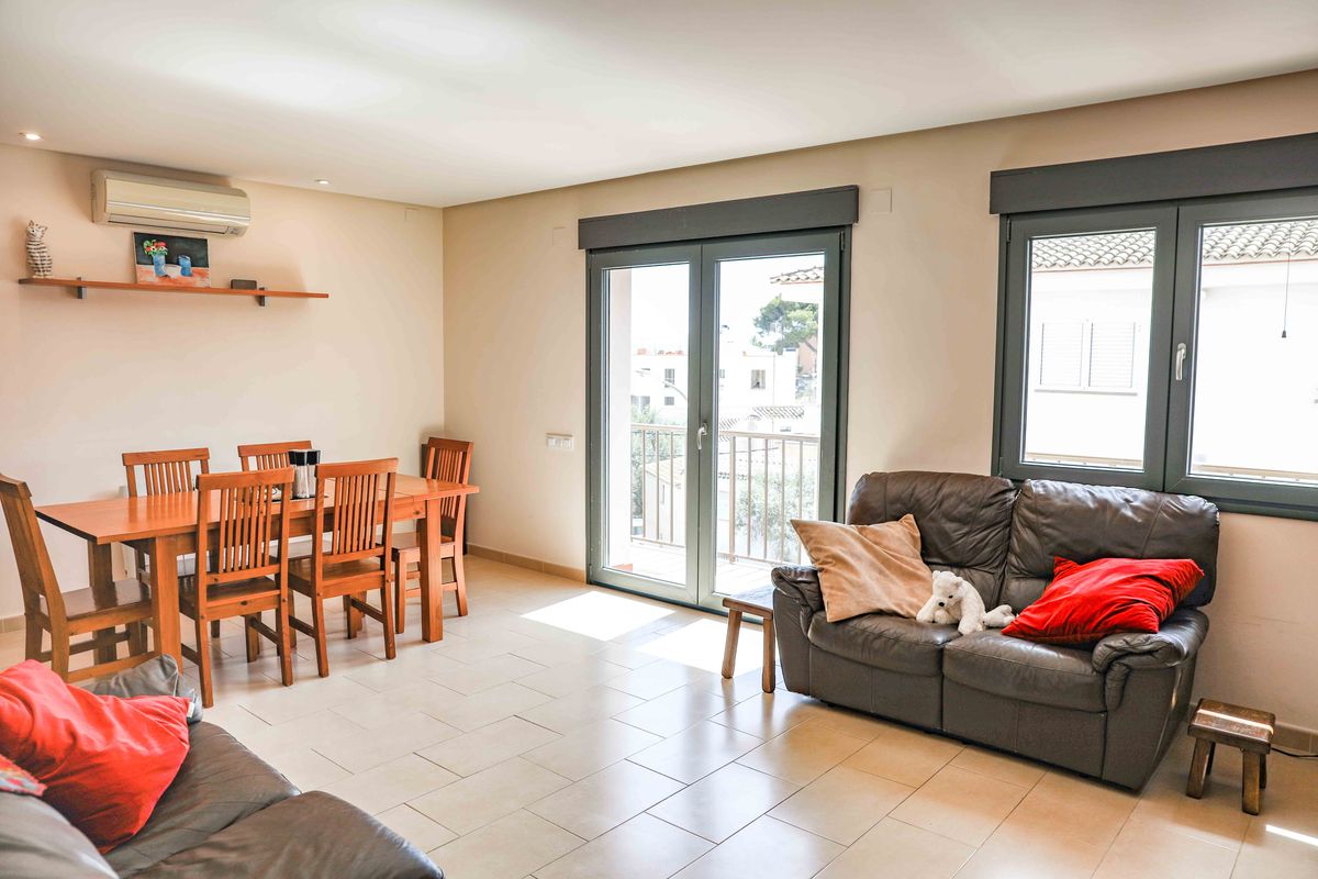 Espace salon-salle à manger lumineux avec sol carrelé, table en bois pour six personnes et canapé en cuir. Grandes portes vitrées ouvrant sur un balcon extérieur.
