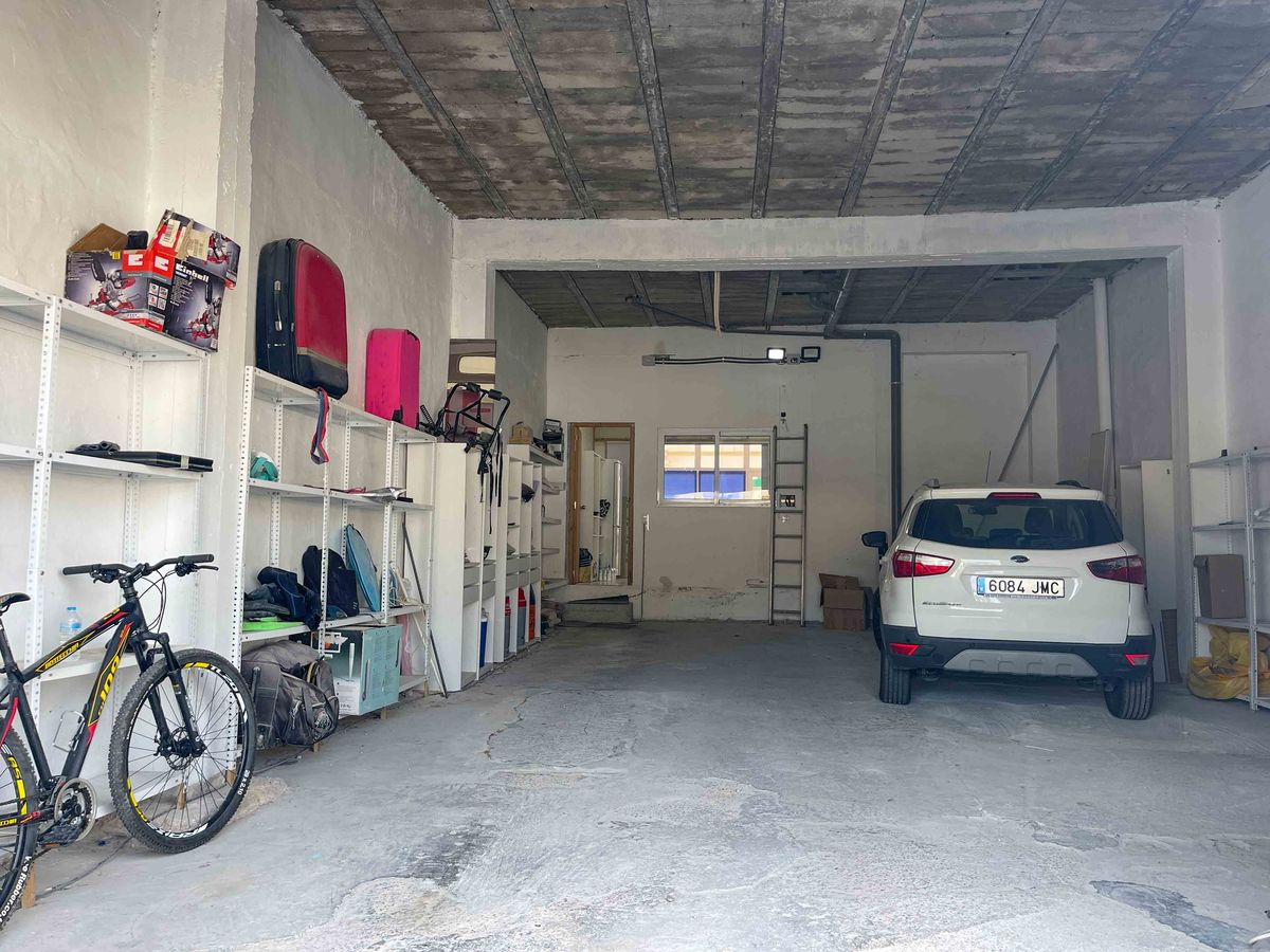 Grand garage avec hauts plafonds en béton et rayonnages métalliques. Espace fonctionnel pour véhicules et stockage bénéficiant d'une fenêtre de ventilation.