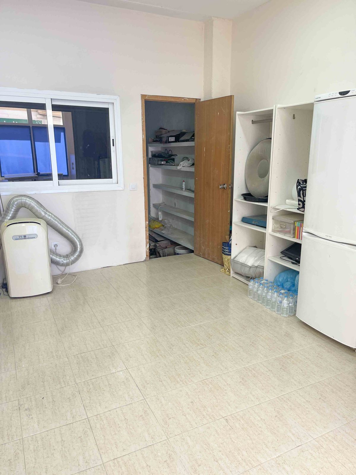 Local technique avec carrelage clair, climatisation portable, étagères de rangement blanches et cellier attenant avec rayonnages.