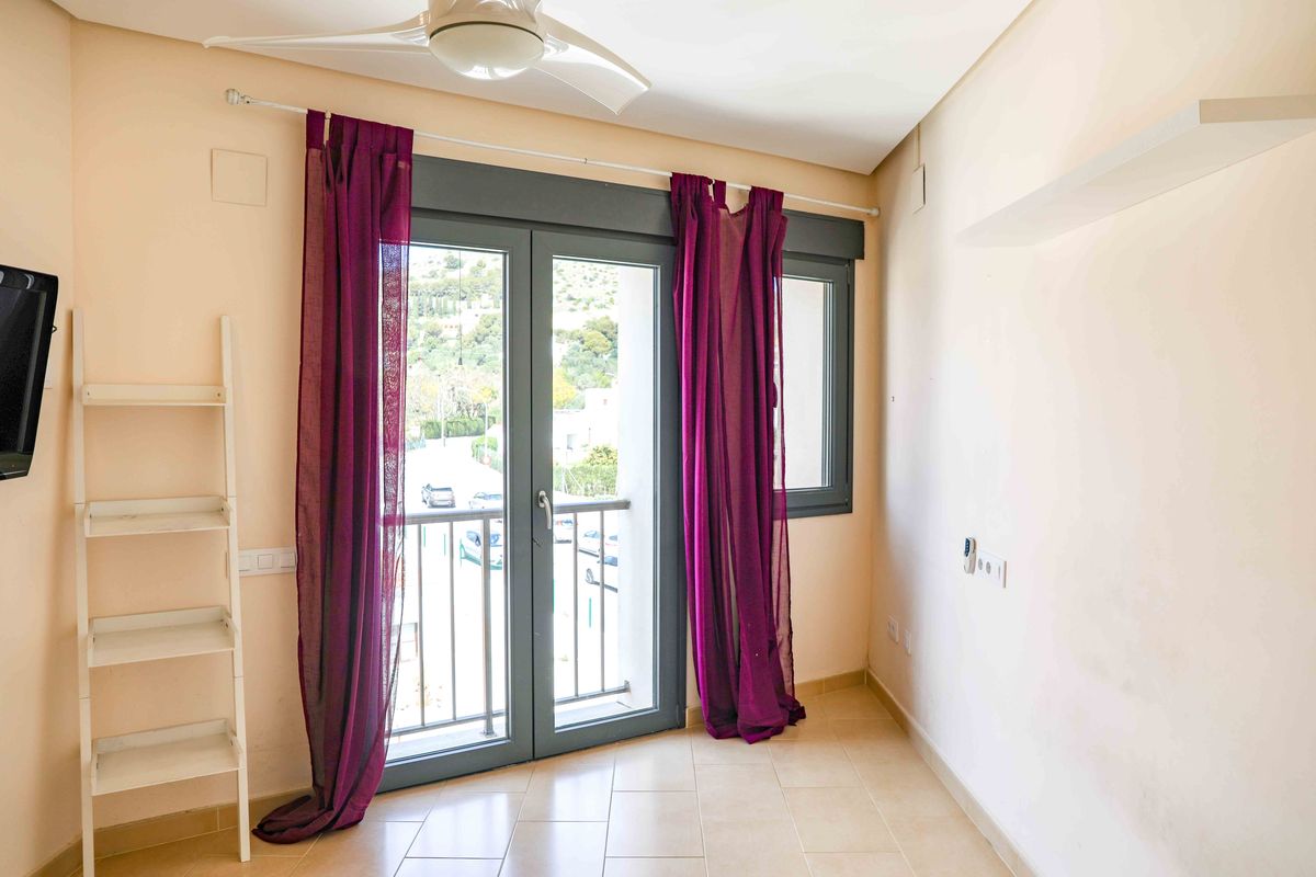 Pièce lumineuse avec portes-fenêtres à cadre sombre, rideaux violets et ventilateur de plafond. Elle dispose d'une étagère échelle blanche et d'un sol en carrelage clair.