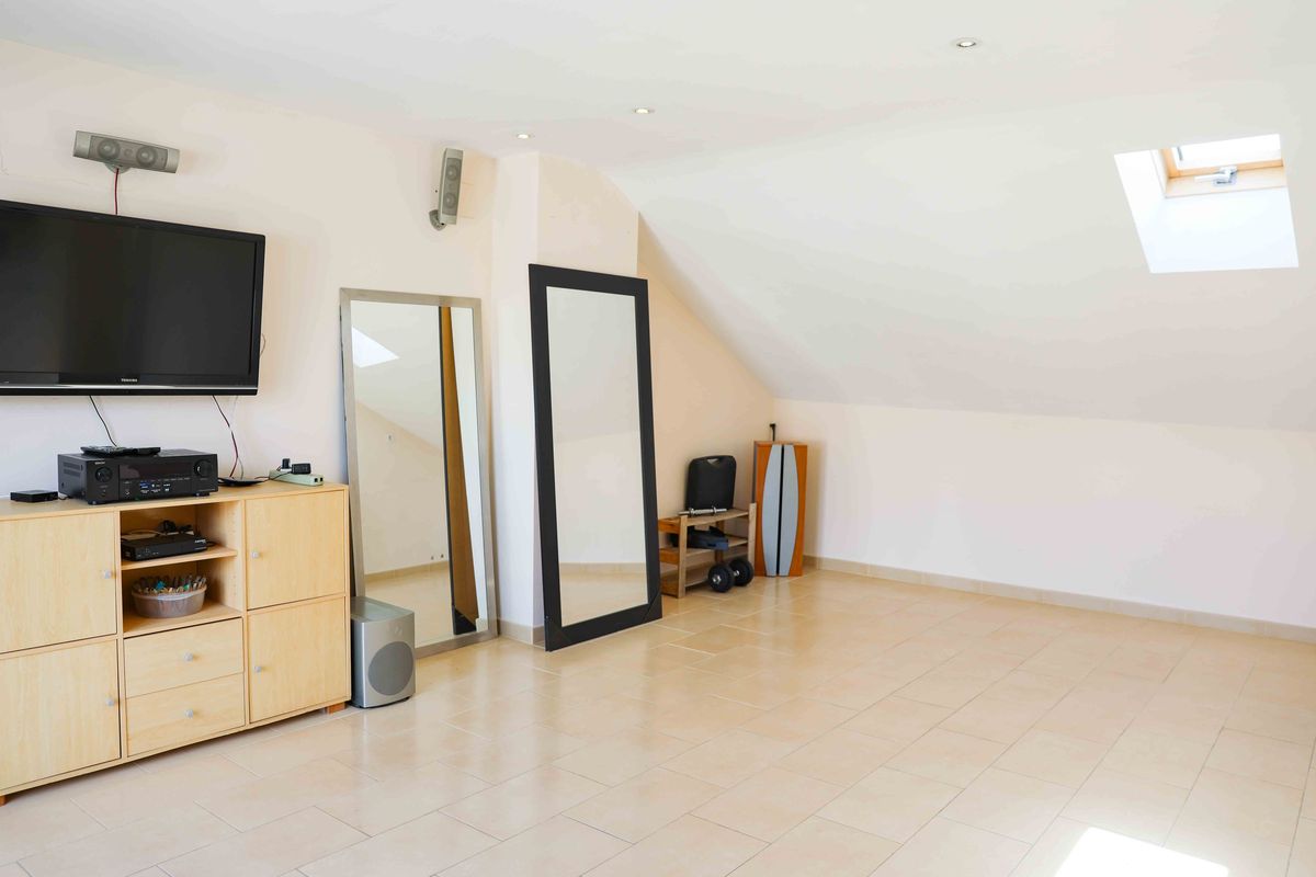 Pièce mansardée lumineuse avec fenêtre de toit, carrelage clair, plafond incliné et espace de divertissement équipé d'un téléviseur et de miroirs.