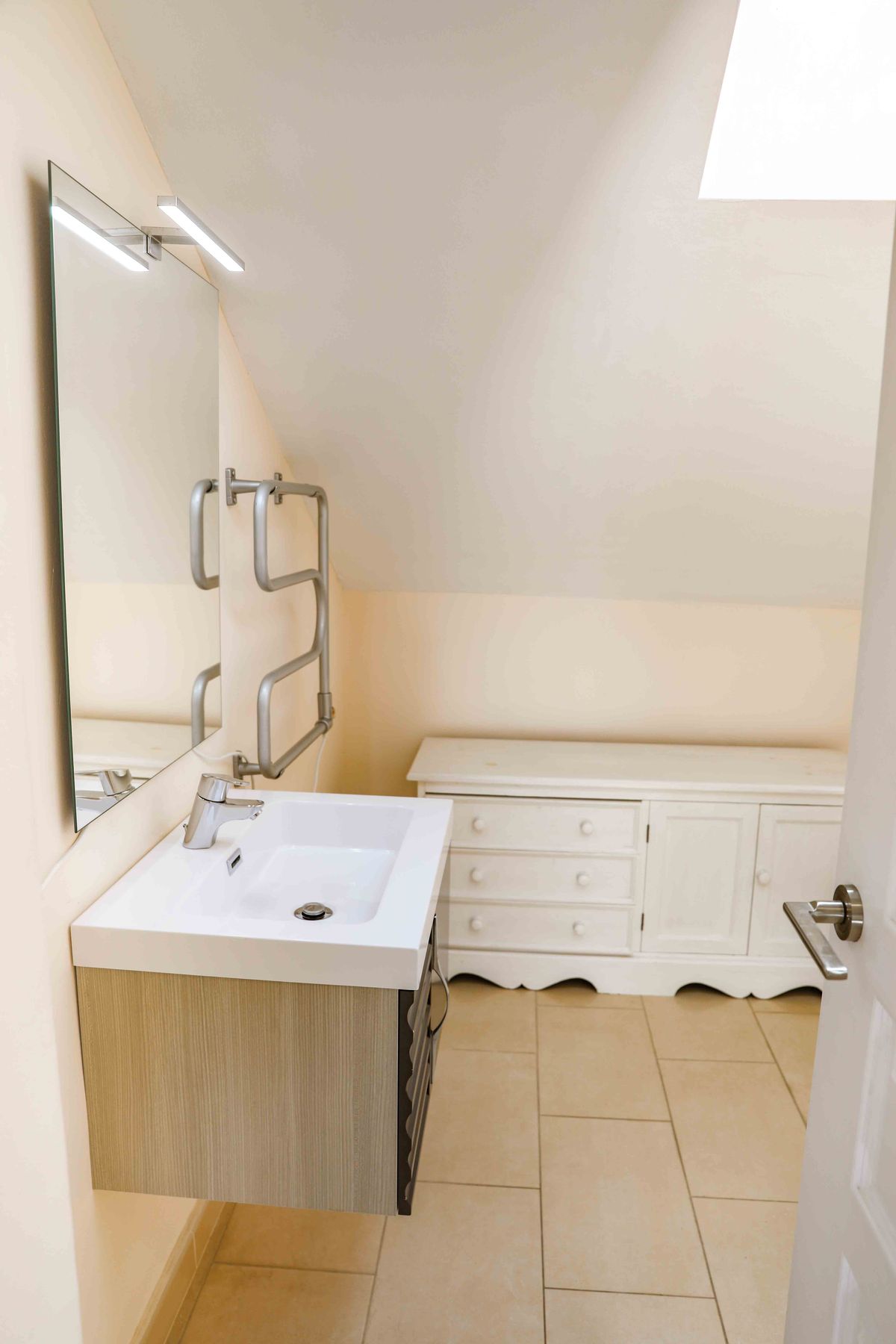 Salle de bain mansardée avec vasque blanche, meuble sous-vasque texturé bois, sèche-serviettes chromé et commode blanche sous plafond incliné.