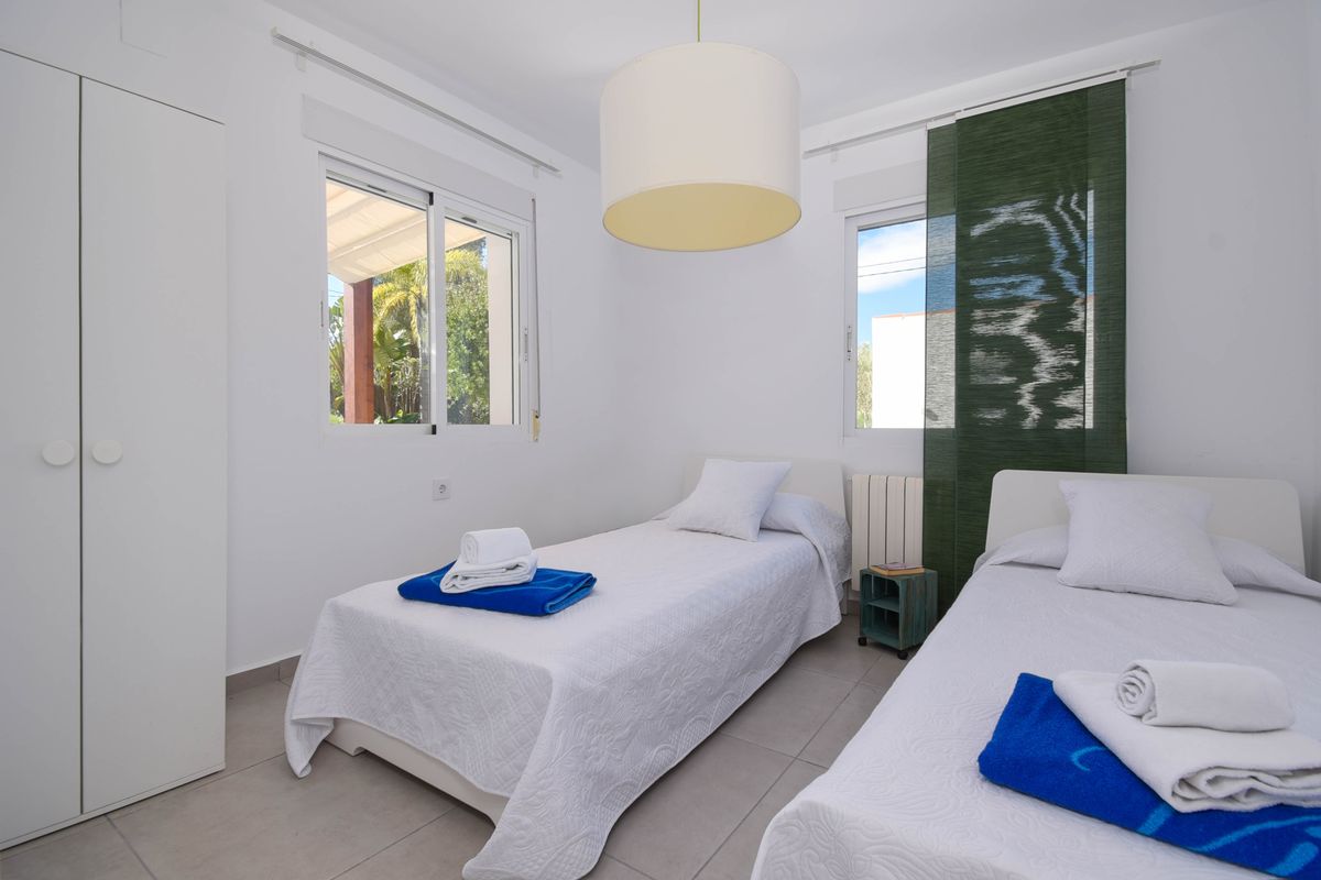 Chambre double lumineuse avec mobilier blanc, armoire minimaliste et fenêtres offrant une lumière naturelle. Dispose d'un sol en carrelage gris et d'une suspension cylindrique.