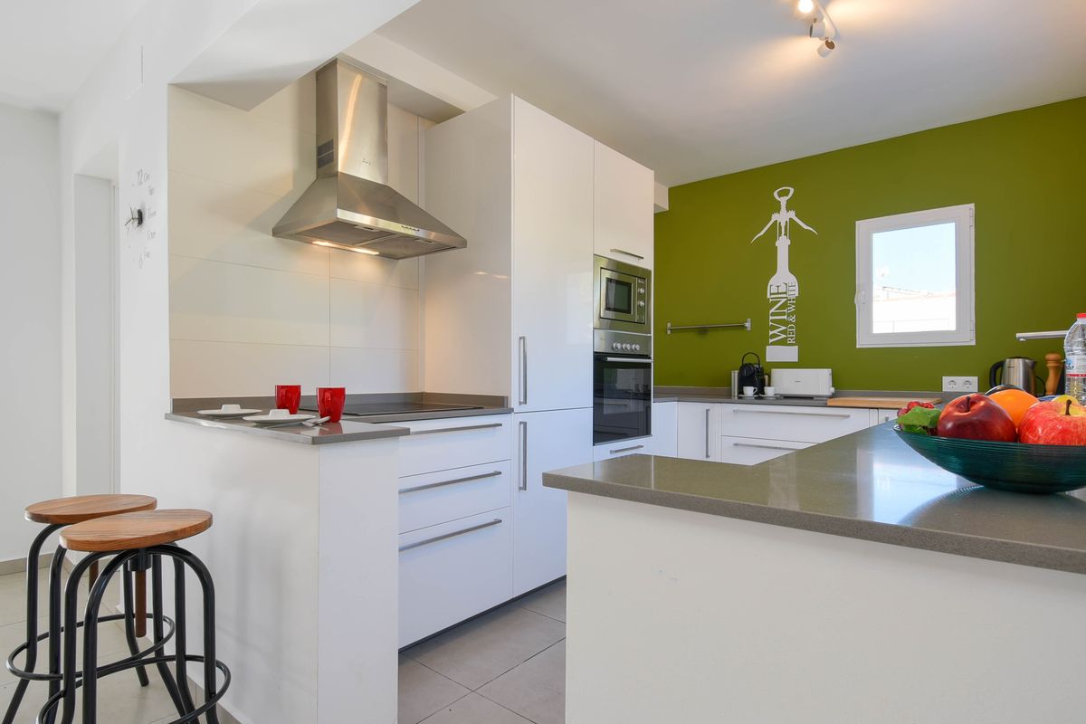 Cuisine moderne avec meubles blancs, plans de travail gris et mur vert. Comprend des appareils intégrés, un bar avec tabourets en bois et une hotte aspirante en inox.