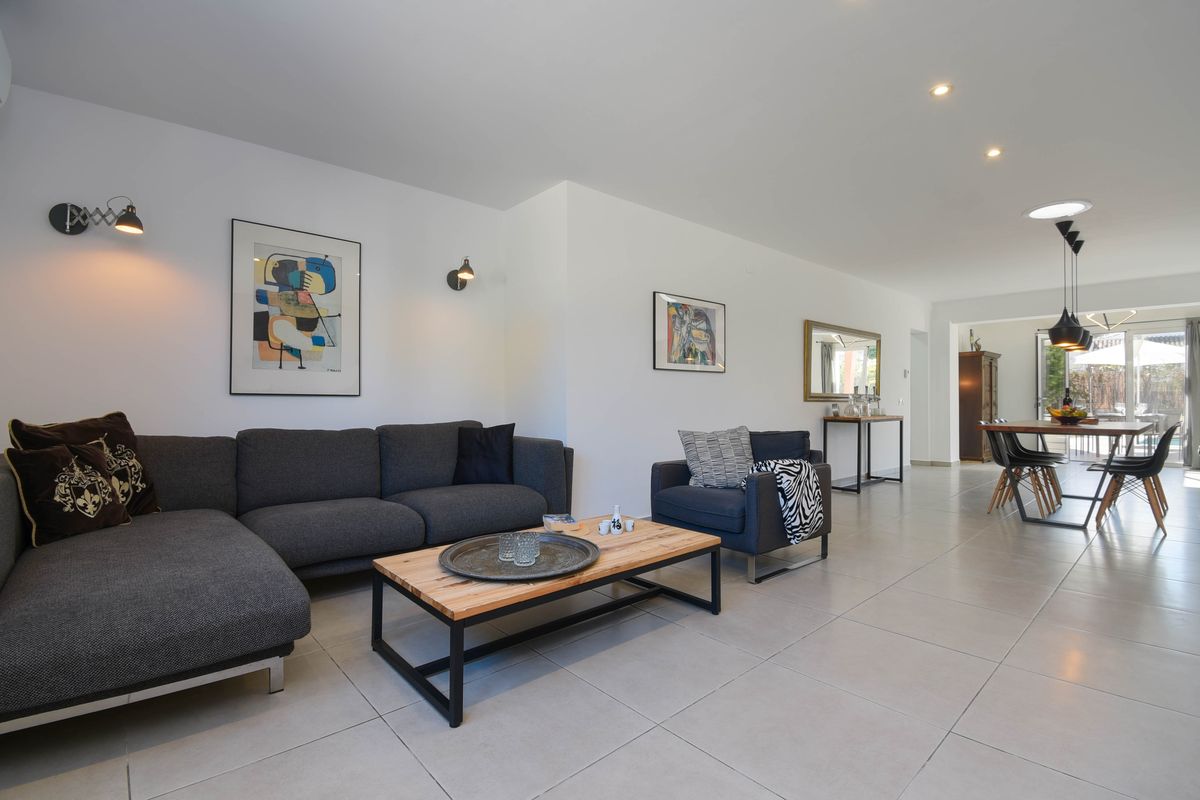 Espace de vie ouvert avec canapé d'angle gris, table basse en bois et métal, et coin repas lumineux menant vers l'extérieur.