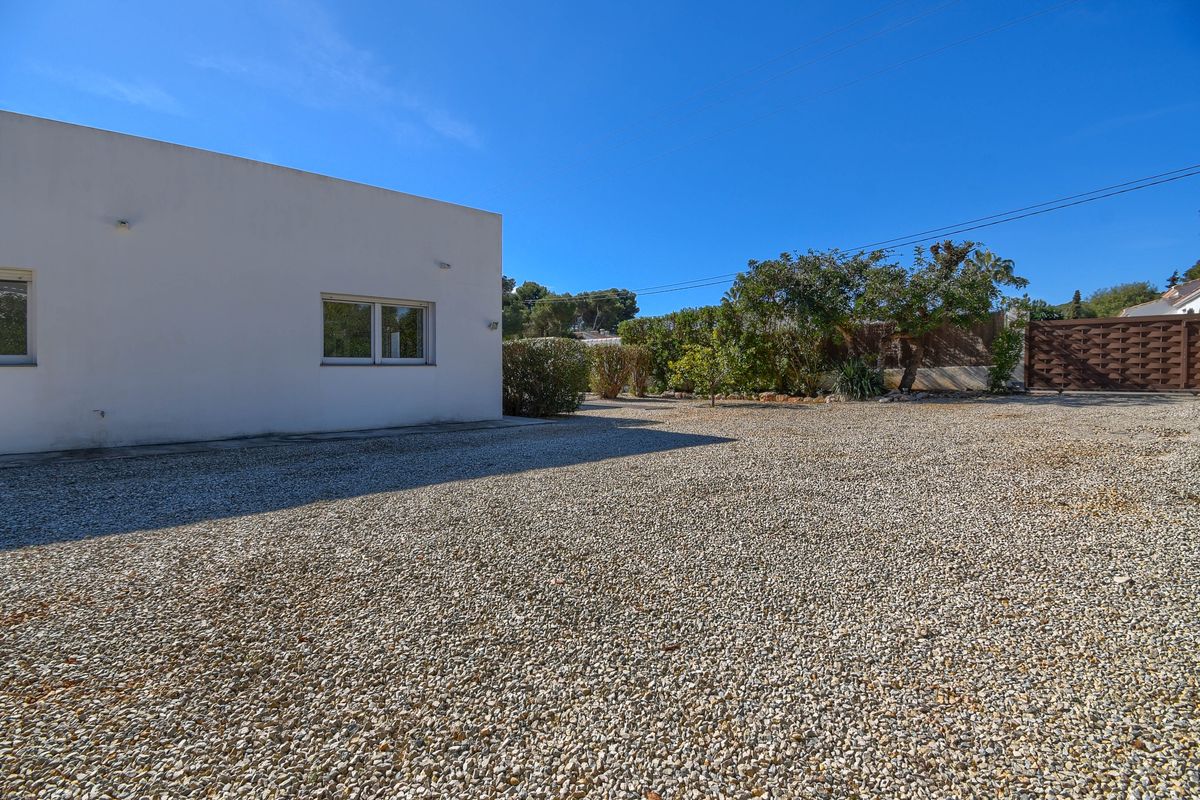 Large allée en gravier longeant une façade blanche épurée, comprenant un portail en bois et une bordure végétale sous un ciel azur.