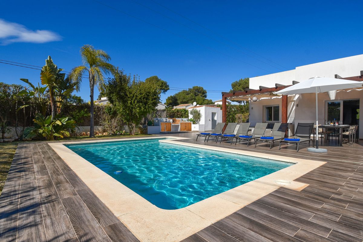 Piscine extérieure avec margelles en pierre, terrasse en carrelage imitation bois, bains de soleil et pergola en bois avec espace repas.