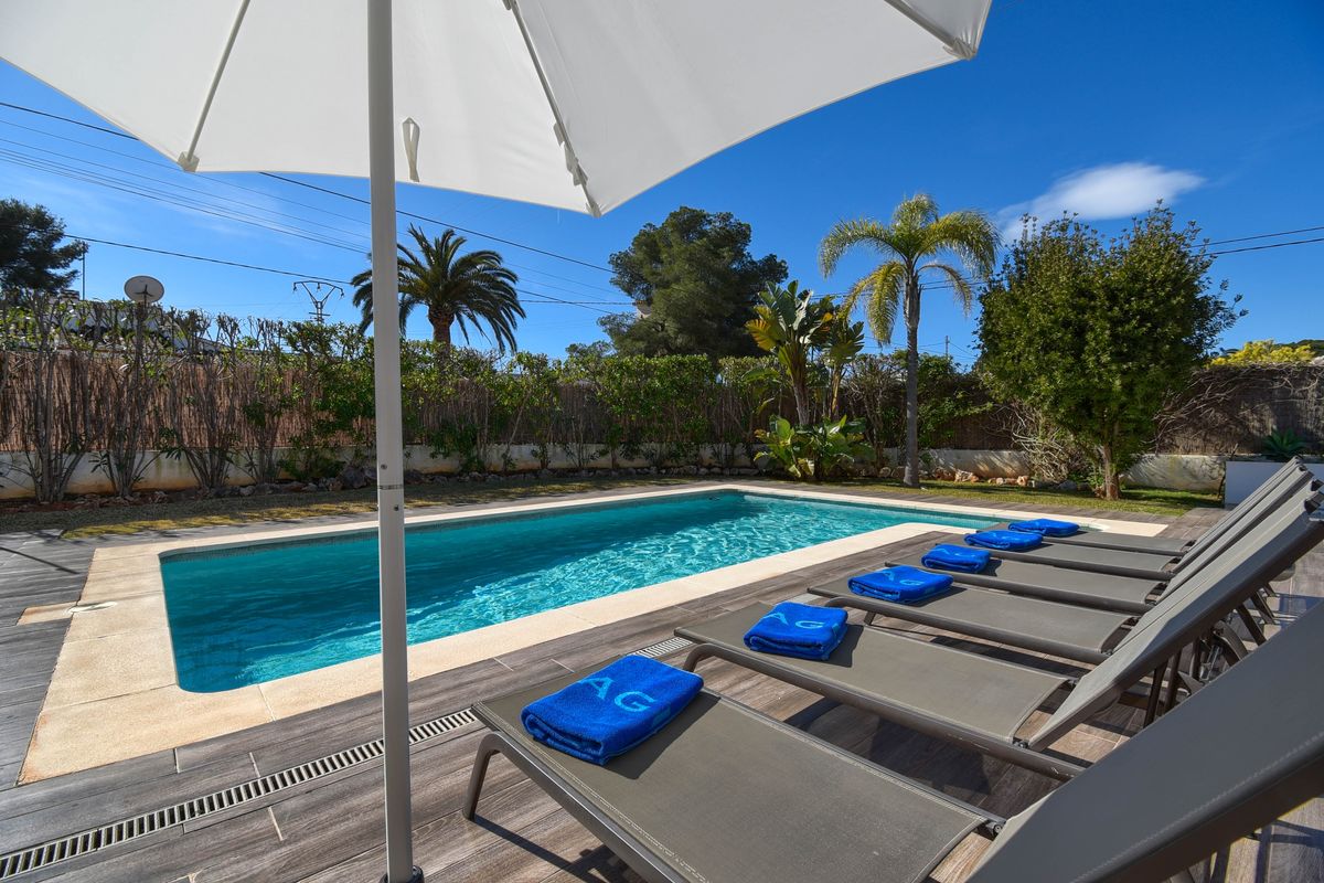 Piscine extérieure rectangulaire avec bordure en pierre, terrasse en grès cérame aspect bois, chaises longues grises et jardin méditerranéen arboré.