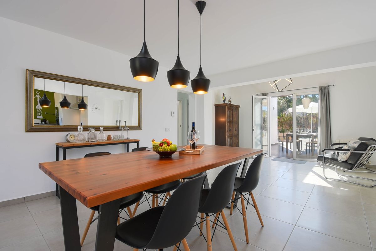 Salle à manger moderne avec table en bois massif, chaises design noires et luminaires suspendus, ouvrant sur une terrasse extérieure lumineuse.