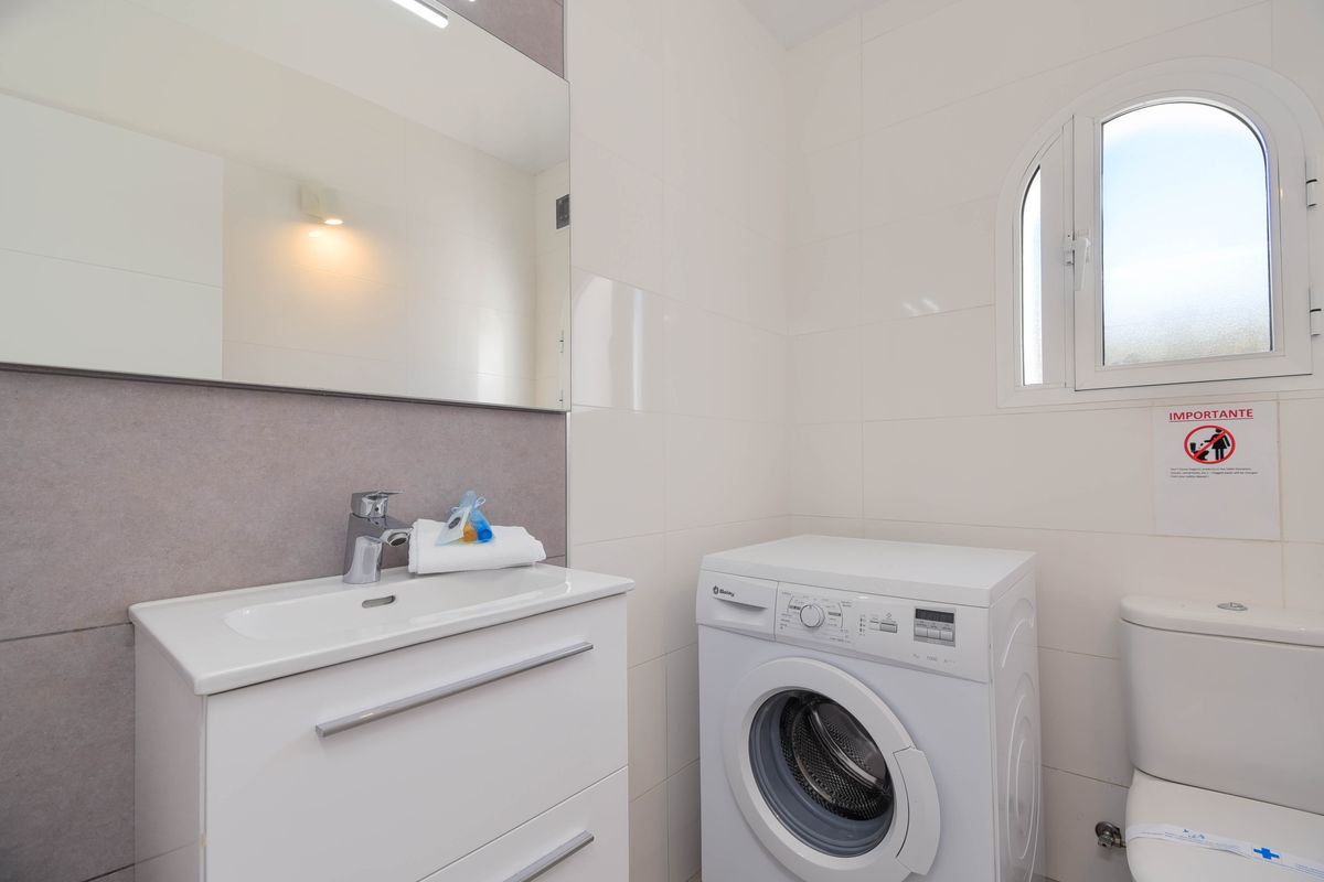Salle de bain fonctionnelle avec meuble vasque blanc, grand miroir et lave-linge. L'espace est doté de carrelage blanc et d'une fenêtre en arc à vitrage dépoli.