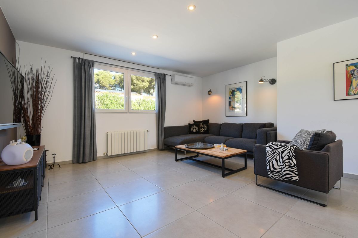 Salon contemporain avec canapé d'angle gris, table basse en bois et carrelage grand format. Équipé de la climatisation et offrant une vue sur la végétation.