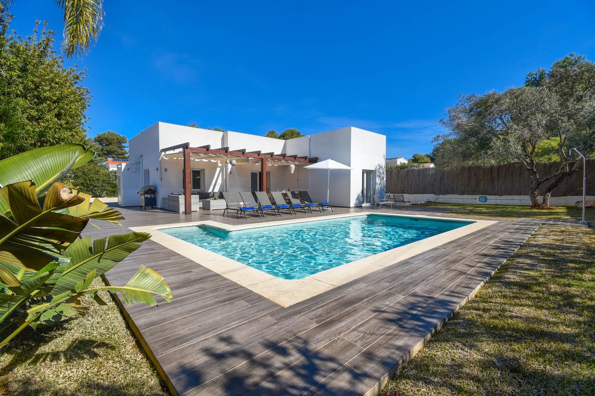 Villa contemporaine blanche avec piscine rectangulaire, terrasse en dalles aspect bois et pergola équipée d'un salon de jardin et de transats.